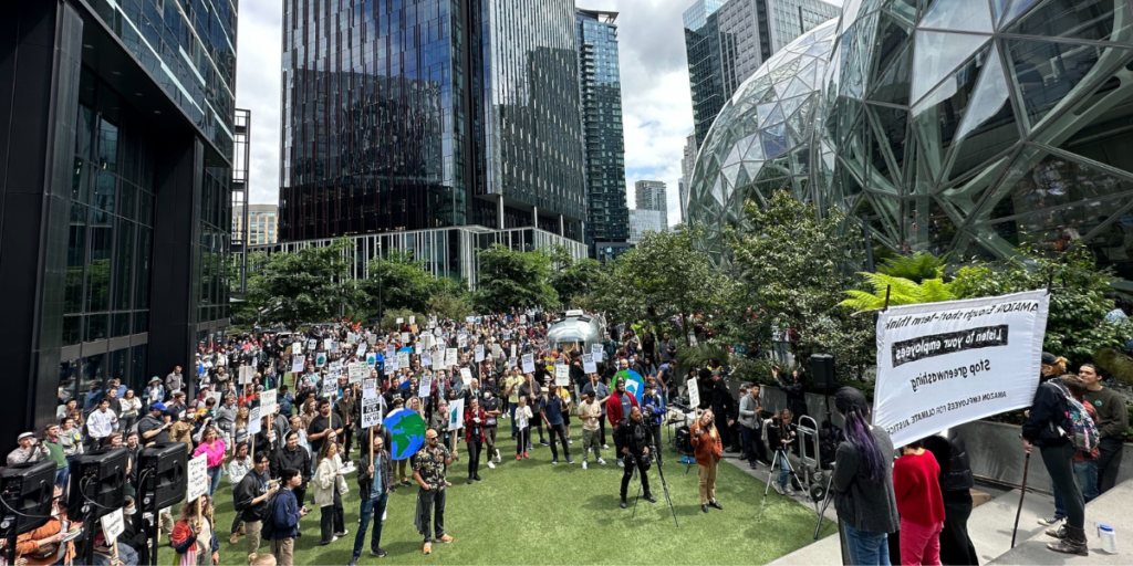 A somber group of Amazon employees gathers outside the company’s Seattle headquarters, with the iconic Amazon logo looming on a glass building behind them, reflecting the recent announcement of 14,000 managerial layoffs in 2025 amid a bustling cityscape.