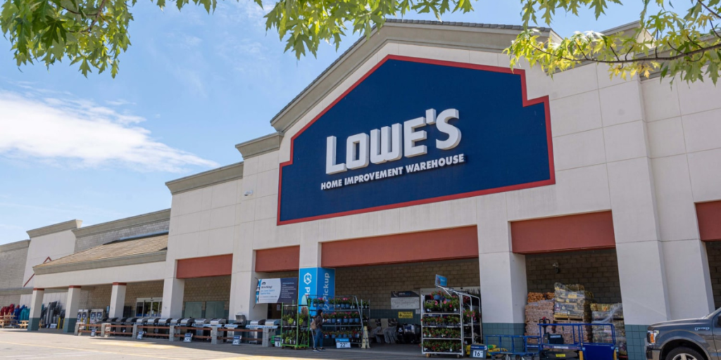 A standard exterior view of a Lowe’s store with its recognizable orange and blue logo, surrounded by a parking lot and clear signage, set against a typical suburban backdrop.