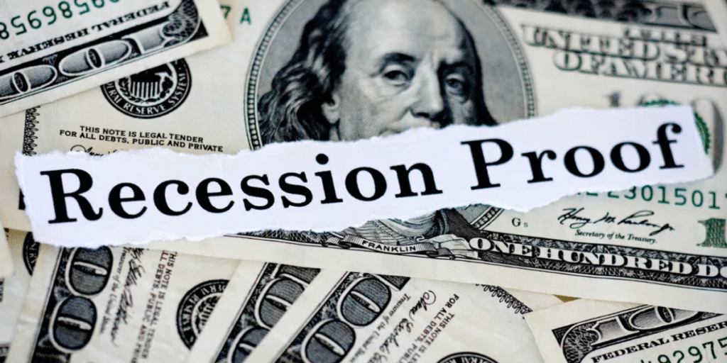 A stack of U.S. dollar bills with the phrase "Recession Proof" boldly printed in gold text across the center, symbolizing financial resilience and strategic planning for 2025. The background features a subtle graph of economic trends, with upward and downward arrows, representing inflation and market volatility.