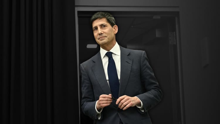 Man in dark suit and navy tie adjusting jacket, standing before a dark background.