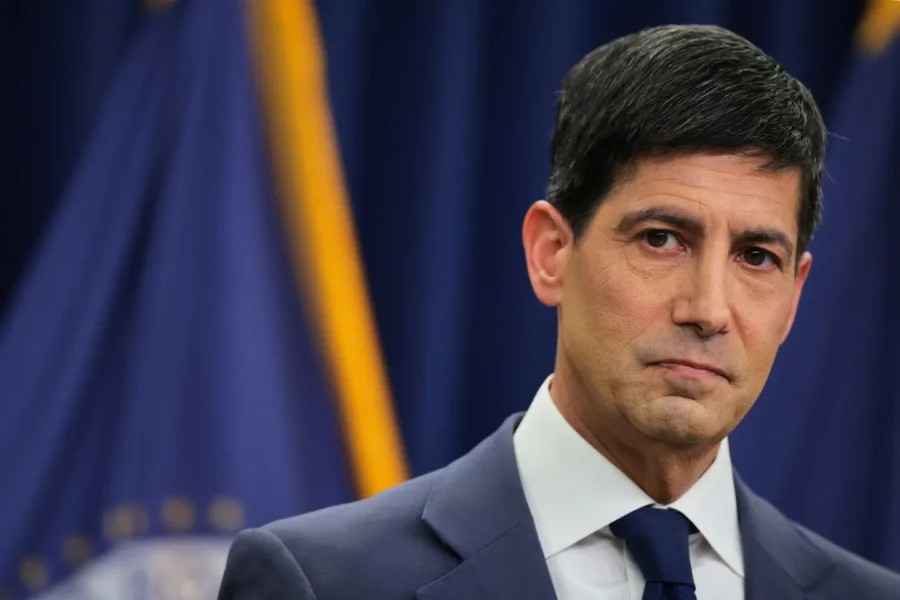 Man in navy suit and blue tie stands before blue drapes and gold flag, with serious expression.
